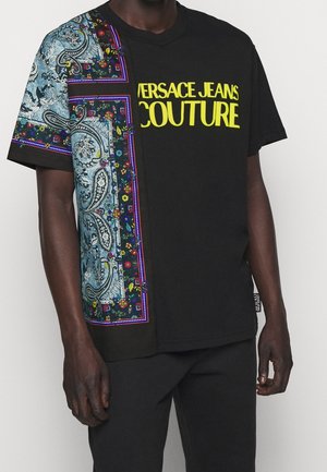 T-shirt noir à manches courtes avec un motif coloré de paisley et floral sur le côté gauche et un texte jaune "Versace Jeans Couture" sur la poitrine.