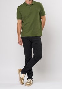 Camisa de polo verde oscuro con cuello y un solo logo, combinada con jeans negros y zapatillas en tonos beige. Conjunto casual en general.