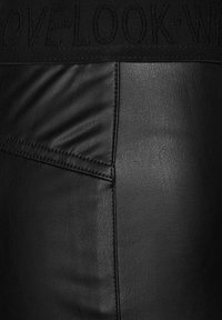 Černé kožené kalhoty s hladkou texturou, viditelnými švy a pasem s vyšitými detaily. Detailní záběr materiálu a designu.