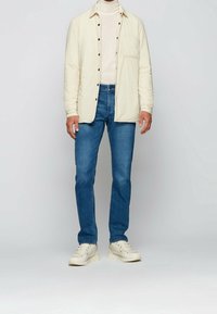 Světle béžová prošívaná košile na knoflíky přes krémový rolák, doplněná modrými džínami a krémovými teniskami.