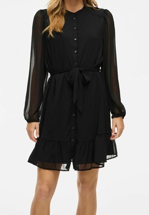 Femme portant une robe noire à boutons avec des manches longues en voile transparent, une ceinture à la taille et un ourlet à volants, posant devant un fond blanc uni.