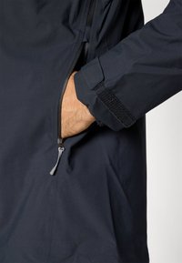 Vandtæt marineblå jakke med lynlåslomme, velcrolukning på ærmet og blød tekstur. Stoffet virker glat.