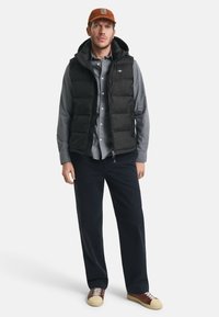 Svart pufferväst med huva, grå skjorta med knappar, marinblå byxor och bruna sneakers med gräddfärgade detaljer. Varm och lager-på-lager-outfit.