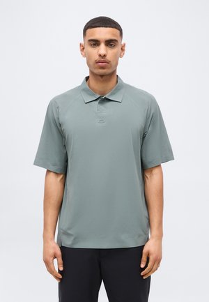 Lühikeste juustega mees, kes kannab tuhmrohelise lühikese varrukaga polosärki ja õrnavärvilisi musti pükse, seisab ühevärvilise hele tausta ees.