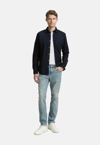 Dunkelblauses Hemd mit weißen Punkten, kombiniert mit hellblauen Jeans und weißen Sneakers, vor einem schlichten weißen Hintergrund stehend.