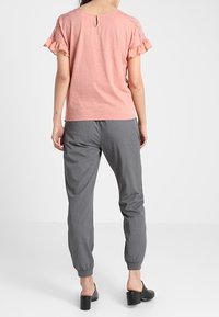 Kvinna klädd i en rosafärgad kortärmad topp med spetsdetaljer och grå ankel-långa byxor, stående vänd bort från kameran mot en vit bakgrund.