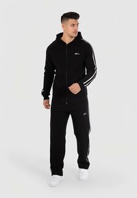 Sort lynlåshættetrøje med hvide sidelister, kombineret med matchende sorte bukser. Har en frontlomme og et logo på brystet.