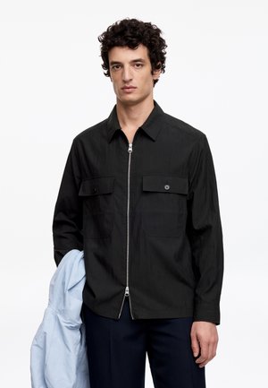 Man met krullend donker haar, die een zwart jack met rits draagt, en een lichtblauw jack in zijn linkerhand houdt, tegen een witte achtergrond.