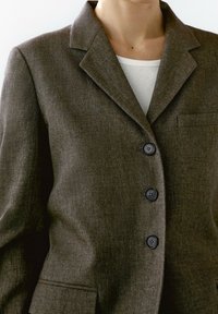 Ruskea räätälöity bleiseri, jossa on lovettu kaulus, kolme etupainiketta ja rintatasku, valmistettu teksturoidusta villasekoitekankaasta.