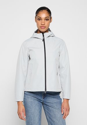 Kobieta w jasnoszarej kurtce z kapturem i czarnym zamkiem oraz niebieskich dżinsach, stojąca na jednolitym jasnym tle.