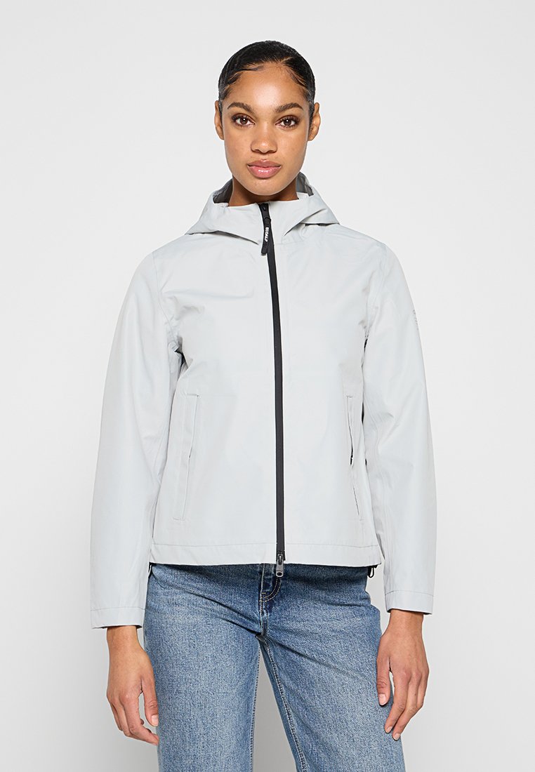 Femme portant une veste à capuche gris clair avec une fermeture éclair noire et un jean bleu, debout devant un fond clair uni.