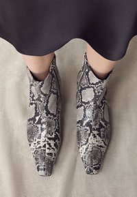 Ghete scurte cu imprimeu piele de șarpe în nuanțe de gri și negru, cu vârf ascuțit și fermoar lateral. Suprafață texturată cu finisaj lucios.