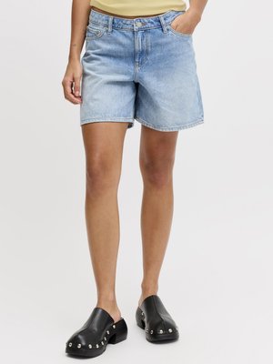 Άτομο που φοράει ανοιχτό μπλε τζιν σορτς, μαύρα παπούτσια χωρίς κορδόνια με καρφιά και ένα κίτρινο μπλουζάκι μαζεμένο μέσα, στέκεται με το ένα χέρι στην τσέπη.