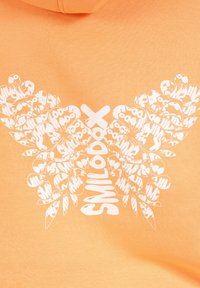 Oranje hoodie met een grote witte vlinderafbeelding, gemaakt van kleinere tekst- en bladvormen, met een zachte, katoenen textuur.