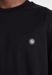 T-shirt noir à col rond avec un petit logo circulaire portant les lettres "B" et "S" sur la poitrine gauche.