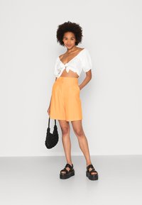 Top cropped branco com mangas bufantes e detalhe em nó, combinado com calções ajustados laranja brilhante. Acessórios incluem uma mala preta texturizada e sandálias de plataforma.