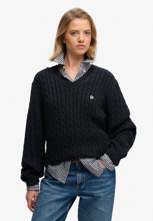 Suéter de punto trenzado en color navy con escote en V, que destaca por su textura prominente y un pequeño logo. Llevado sobre una camisa de rayas blanca y navy.