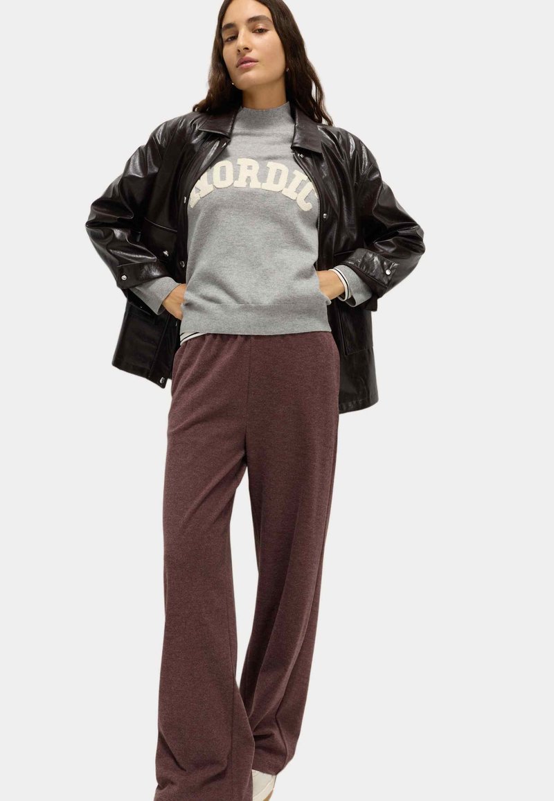 Zwarte glanzende oversized jas, grijze trui met de tekst "NORDIC" en losse kastanjebruine broek. Texturen contrasteren tussen de verschillende stoffen.