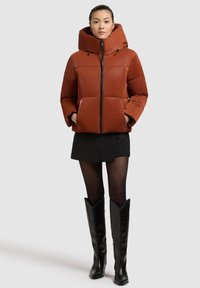 Pomarańczowa kurtka puchowa z wysokim kołnierzem, zamkiem z przodu i dużym kapturem. Para czarnych kozaków do kolan dopełnia stylizację.