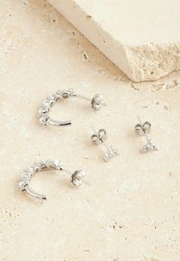 Silberne Hohrringe mit kreisförmigen Akzenten und zwei funkelnden Steckerohrringen. Glatte Textur mit glänzendem Finish, auf einem beigen Untergrund angeordnet.
