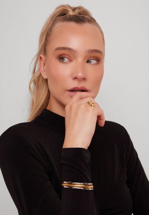 Bague en or avec un design texturé portée à la main, accompagnée d'un bracelet en or tressé. Le modèle a une queue de cheval et porte un haut noir.