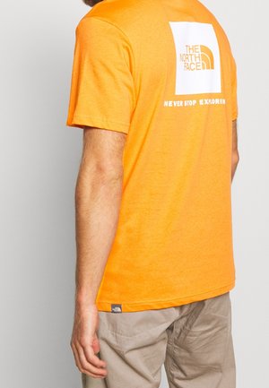Mężczyzna w jaskrawo pomarańczowej koszulce The North Face z białym logo i napisem "Never Stop Exploring" na plecach, w połączeniu z beżowymi spodniami.