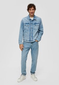 Jasnoniebieska kurtka dżinsowa z dwoma kieszeniami na piersi, z zapięciem na guziki i lekko przetartym wykończeniem, noszona na pasujących jasnoniebieskich dżinsach i białych trampkach.