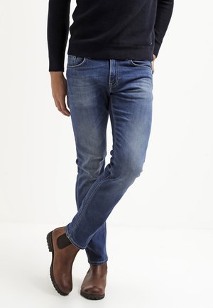 Mann trägt blaue Jeans, braune Chelsea-Stiefel aus Leder und einen schwarzen Pullover, steht mit gekreuzten Beinen vor einem weißen Hintergrund.