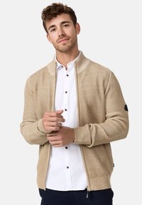 Cárdigan de punto beige con cremallera, textura acanalada, cuello alto y parche negro con logotipo en la manga izquierda. Combinado con una camisa blanca de botones.