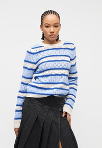 Νεαρή γυναίκα με πλεξούδες, φοράει πλεκτό πουλόβερ με μπλε και άσπρες ρίγες και μαύρη πλισέ φούστα με σκίσιμο μπροστά.