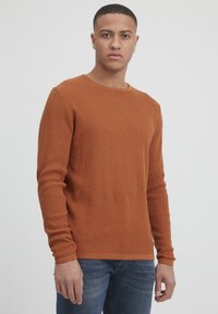 Pullover din bumbac de culoare portocalie arsă, cu mâneci lungi și un guler rotund, stilizat cu blugi, purtat pe un fundal neutru.