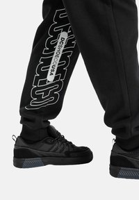 Schwarze Sportschuhe mit strukturiertem Obermaterial, das ein elegantes Design und eine Gummisohle aufweist, kombiniert mit schwarzen Jogginghosen mit weißem Branding.