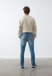 Suéter beige, jeans de mezclilla azul, corte ajustado, costuras visibles, vista trasera, diseño casual, zapatillas negras con suelas blancas.