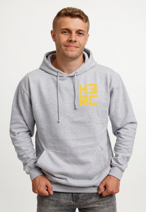 Ung mand iført lysegrå hoodie med gul "HERO" tekst foran, står mod en ensfarvet hvid baggrund med hænderne nær lommerne.