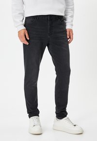 Musta farkkuhousut, joissa on slim fit -leikkaus, viisi taskua ja hillityt kulutukset. Yhdistetty valkoisiin tenni- tai lenkkareihin ja vaaleanväriseen huppariin.