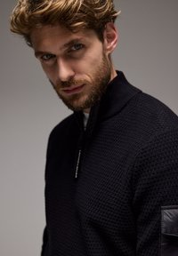 Svart stickad tröja med dragkedja i halsen, med ett strukturerat mönster och ett slätt panel på axeln. Modellen visar ett neutralt uttryck.