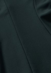 Dunkelgrüner Stoff mit glatter Textur, der einen leichten Glanz aufweist und mit gefalteten Faltendetails versehen ist, die durch subtile Nähte angedeutet werden.