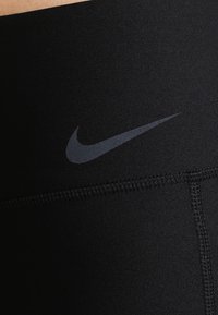 Musta urheiluhousut joustavasta kankaasta, jossa on sileä pinta, ja vyötäröltä löytyy tummanharmaa Nike-logoswush.