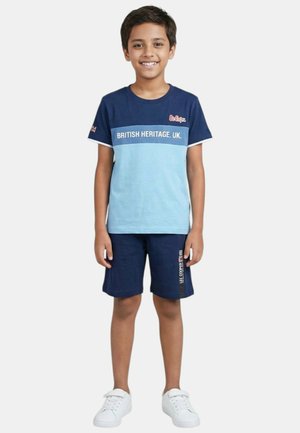 Garçon debout, portant un t-shirt bleu "British Heritage. UK.", un short marine avec texte, et des baskets blanches, souriant sur un fond blanc.