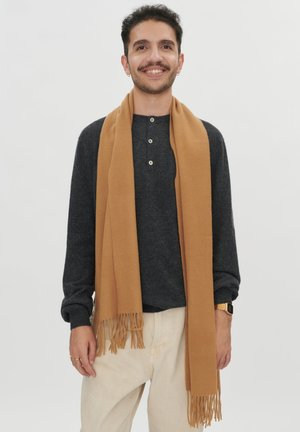 Lächelnder Mann mit dunklem lockigem Haar trägt goldene Creolen, einen grauen, geknöpften Pullover, beige Hose, einen Kamelhaarschal und eine goldene Uhr, steht vor weißem Hintergrund.