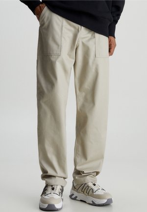 Man draagt beige rechte broek met voorzakken en witte chunky sneakers, staat met één hand in de zak.