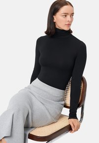 Top negro de manga larga con cuello alto, combinado con una falda gris claro acanalada hasta la rodilla. Modelo sentado en una silla de madera y metal.
