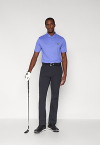 Svetlo vijolična polo majica s ovratnikom in tremi gumbi, črne golf hlače, črni čevlji, drži golf palico, nosi belo rokavico.