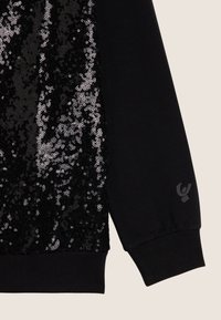 Sudadera negra con un panel frontal cubierto de lentejuelas. Los puños y el dobladillo son de canalé, con un pequeño detalle de logo cerca de la muñeca.