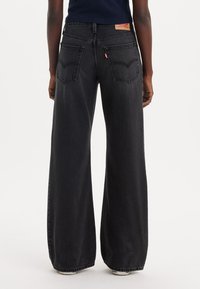 Laajalti leveät mustat farkut, jotka on valmistettu denimistä. Niissä on kaksi takataskua, hienovaraiset ompeluyksityiskohdat ja vaaleanruskea merkkilappu vyötäröllä.