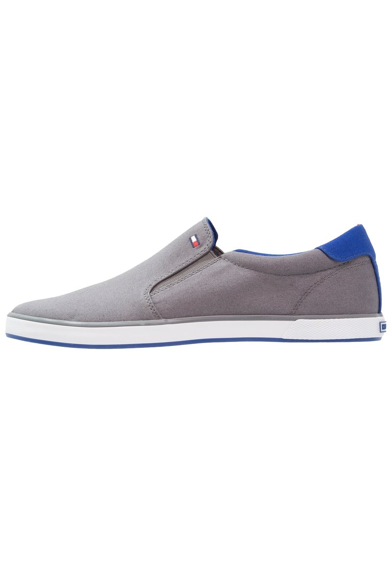 Scarpa slip-on grigia con un accento blu sul tallone, realizzata in canvas, con una suola in gomma bianca e elementi di design minimalisti.
