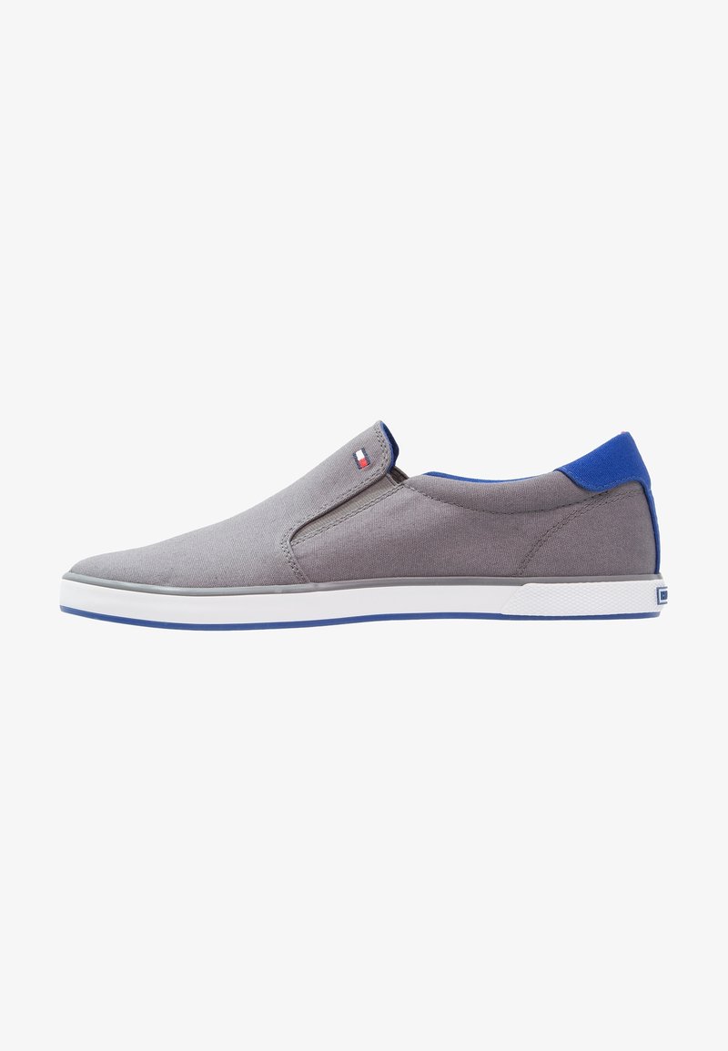 Scarpa slip-on grigia con un accento blu sul tallone, realizzata in canvas, con una suola in gomma bianca e elementi di design minimalisti.