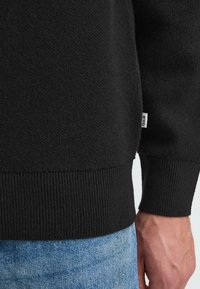 Svart strikket genser med ribbestrikkede mansjetter og nederkant, har en teksturert overflate og en liten merkeetikett på siden. Brukt sammen med lyseblå jeans.