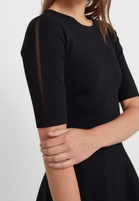 Abito nero a costine con maniche corte, dotato di scollatura a barca e dettagli a righe trasparenti lungo le maniche. Tessuto liscio e elasticizzato.