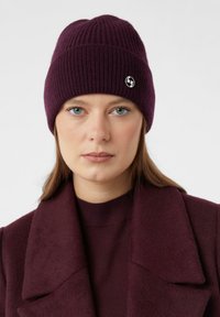 Beanie lavorato a maglia di un viola scuro con texture a coste e un piccolo logo in evidenza. Abbinato a un maglione coordinato di un viola scuro.